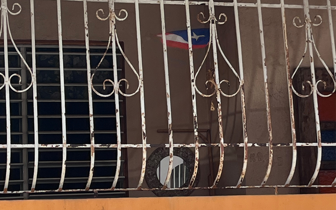 An image of a Puerto Rican flag mounted on a patio. The patio has colorful concrete details and wrought iron fencing that has been painted white.