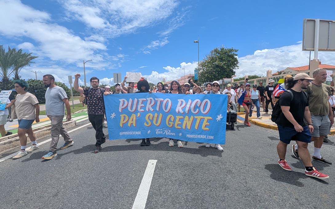 A large group of people march down the street. A group of them carry a sign that reads "Puerto Rico Pa' Su Gente"